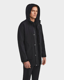 A man in the Rudsak IAN - 6124009 Black rainwear jacket stands slightly turned against a light gray backdrop. The water-repellent coat features snap buttons, pockets, and an upturned hood.
