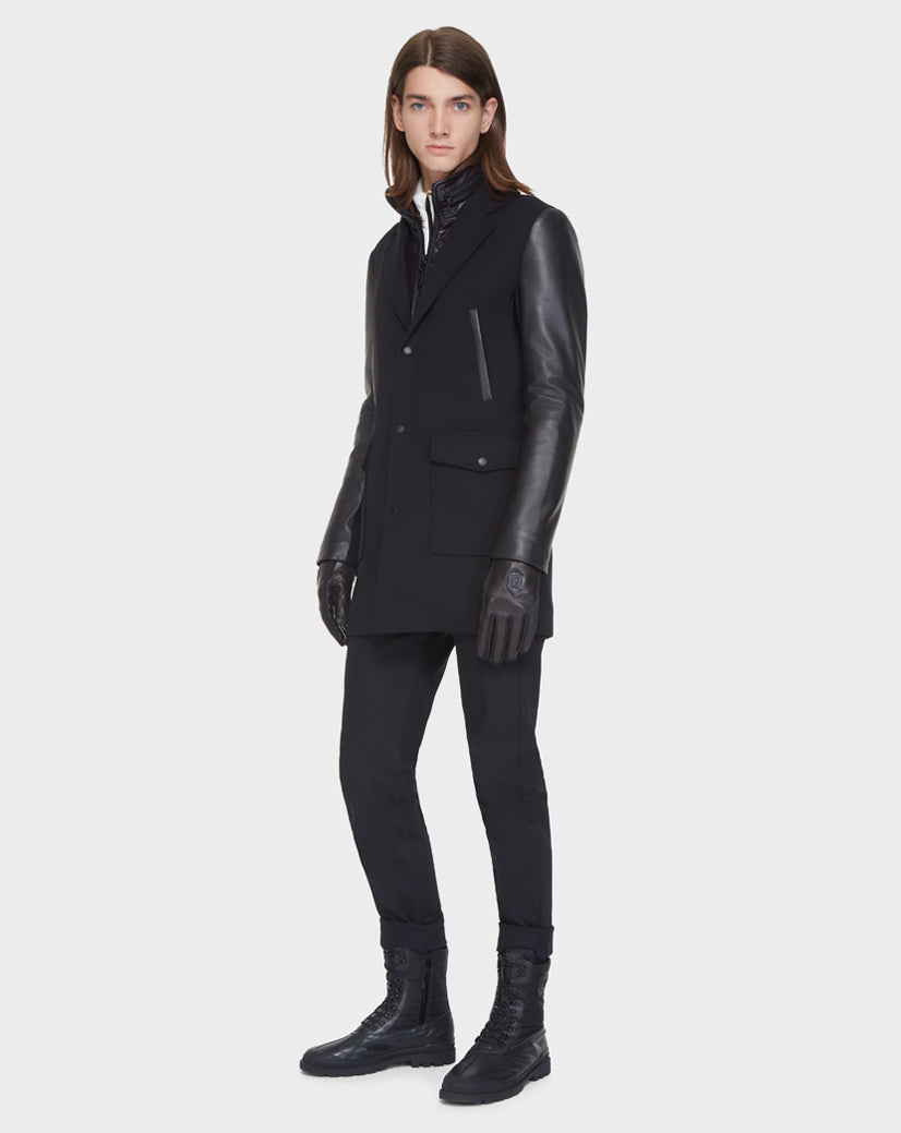 A person with long brown hair wears the Rudsak BEAU - M121504 BLACK men’s coat, featuring leather sleeves, paired with black pants, gloves, and boots against a plain light background.