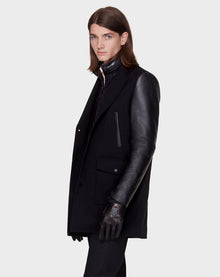A person with long brown hair models the Rudsak BEAU - M121504 BLACK men’s coat with leather sleeves, black gloves, and a zip-up inner layer, standing against a plain light gray background and looking slightly to the side.