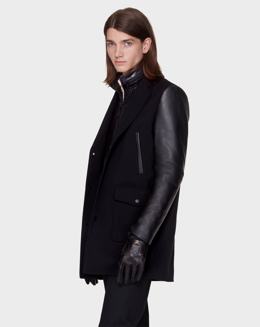A person with long brown hair models the Rudsak BEAU - M121504 BLACK men’s coat with leather sleeves, black gloves, and a zip-up inner layer, standing against a plain light gray background and looking slightly to the side.