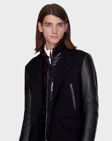 A young person with straight brown hair models the Rudsak BEAU - M121504 BLACK men’s coat with leather sleeves over a glossy black zip-up jacket, standing against a plain light background.