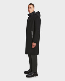 A man stands sideways against a plain white background, wearing the Rudsak DRAL - 6124503 Black mens wool jacket with a hood, paired with dark trousers and black boots.