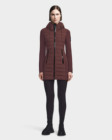 A woman faces forward against a plain white background, wearing RUDSAK's MERINA - 8122546 CHOCOLATE long brown quilted down jacket with a hood, paired with black leggings and black boots.