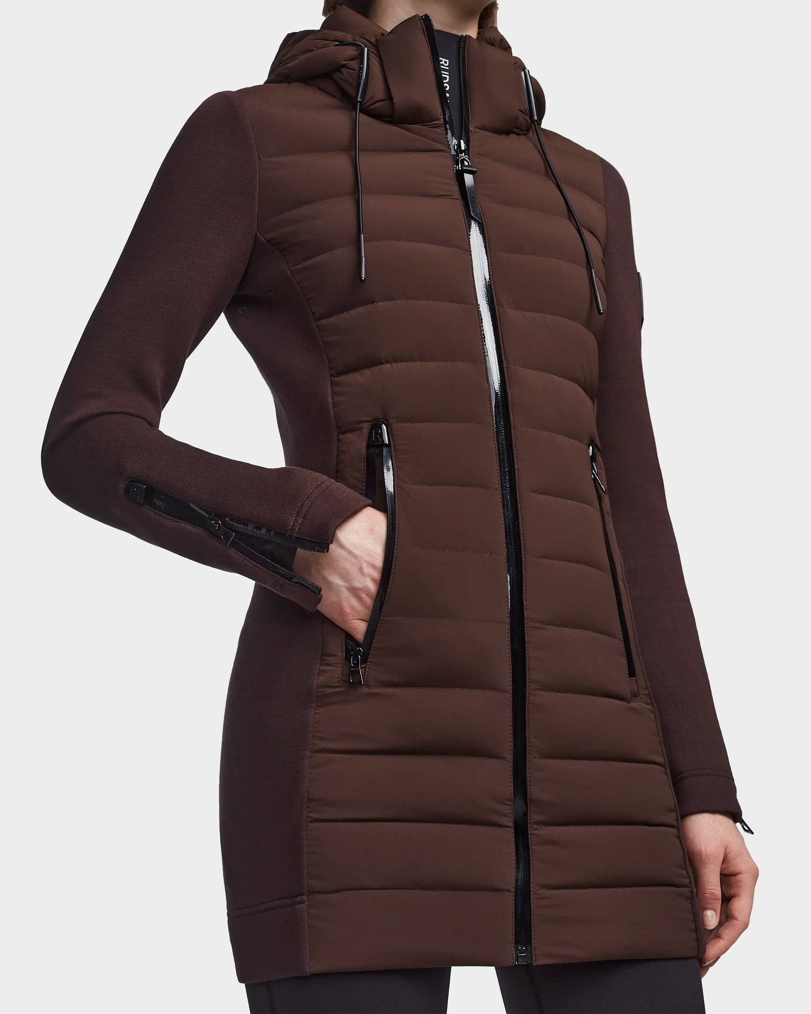 Wearing the RUDSAK MERINA - 8122546 CHOCOLATE quilted down jacket, a person showcases its long brown design with a hood, black zipper details, zippered pockets and cuffs, and fitted sleeves—perfect as a transitional piece with one hand in a front pocket.