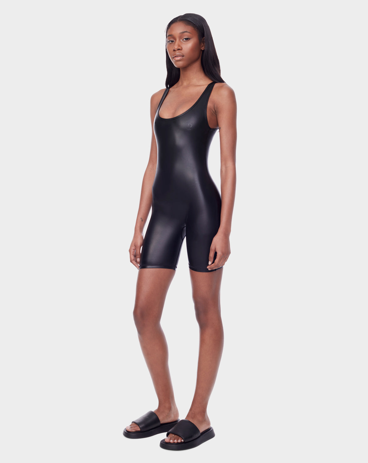 A woman wears the Rudsak AVERI - 8523015 BLACK fitted, shiny, sleeveless romper with shorts and black slide sandals against a plain white background.