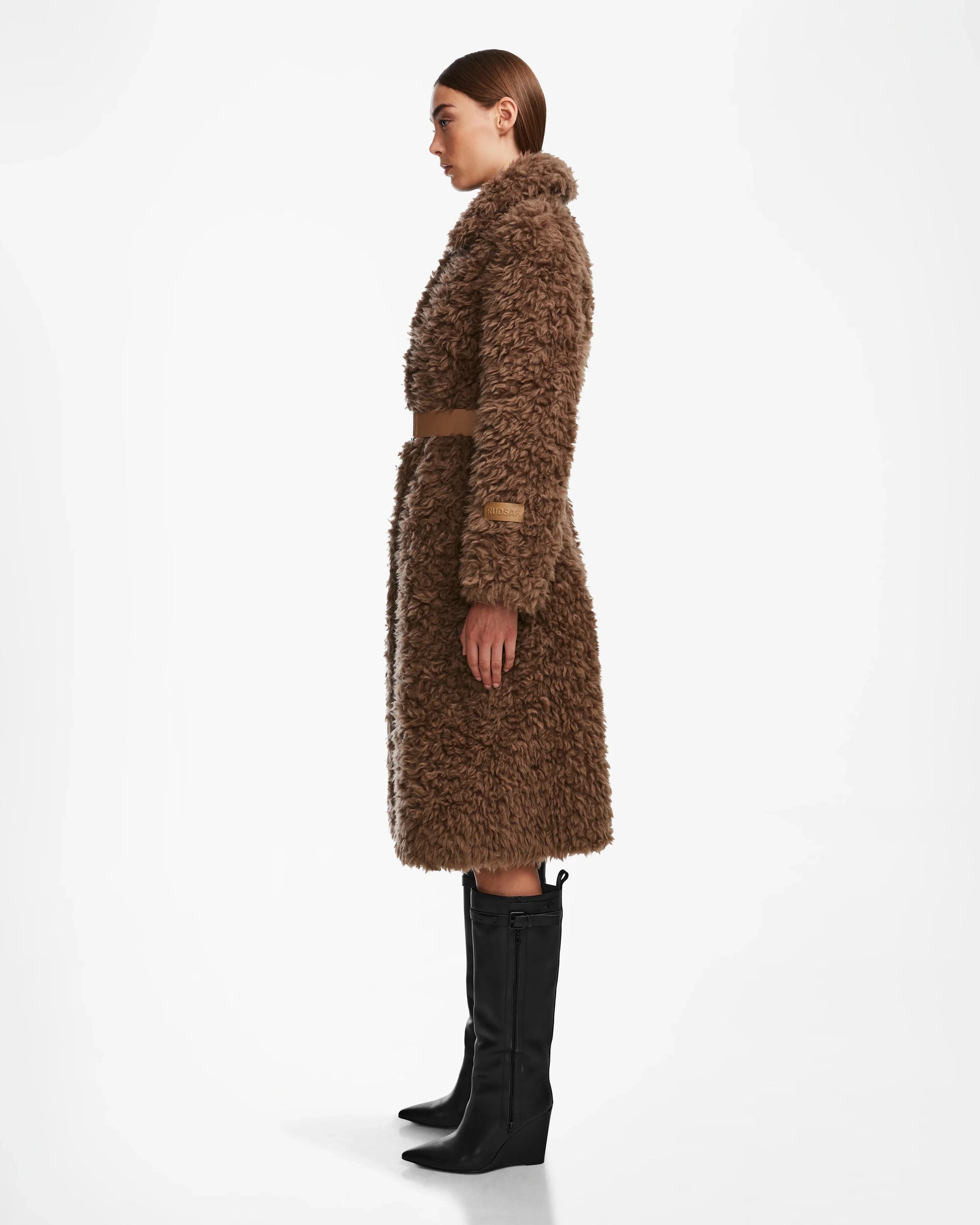 A woman stands in profile wearing the KAMILAH - 8124827 Camel faux fur trench coat by Rudsak with a vegan leather belt, paired with black knee-high boots, against a plain white background.