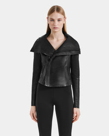 A woman with straight blonde hair wears the Rudsak AVRIL - 8124561 Black leather jacket, featuring a wide collar, paired with black fitted pants, standing against a plain light gray background.