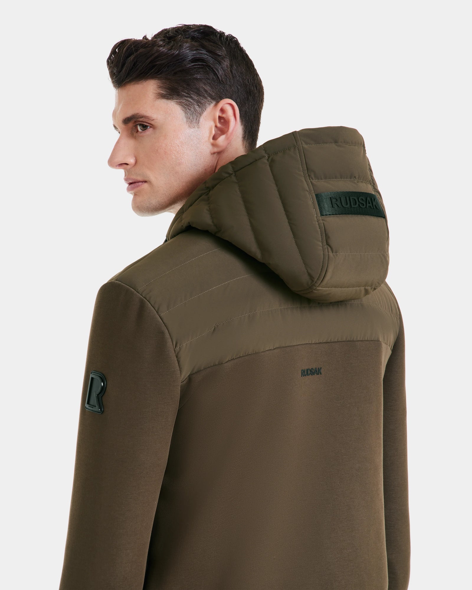 A man wears the DEE MAT - 6124028 Army, an olive green Rudsak mens lightweight hybrid down jacket with RUDSAK branding on the hood and back, looking over his shoulder against a plain white background.