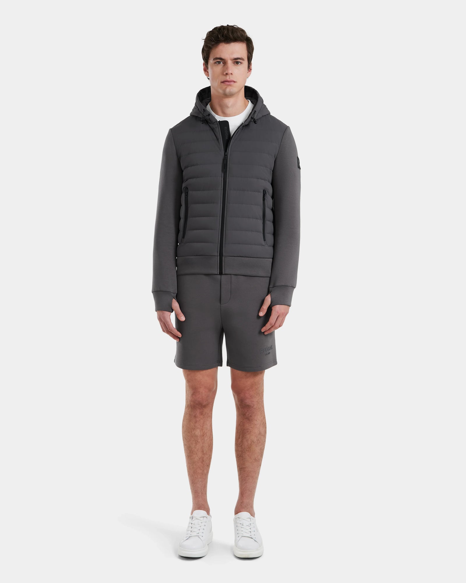 A young man faces forward against a plain white background, wearing the Rudsak DEE MAT - 6124028 Grey men’s lightweight down jacket, paired with matching grey shorts, a white t-shirt, and white sneakers.