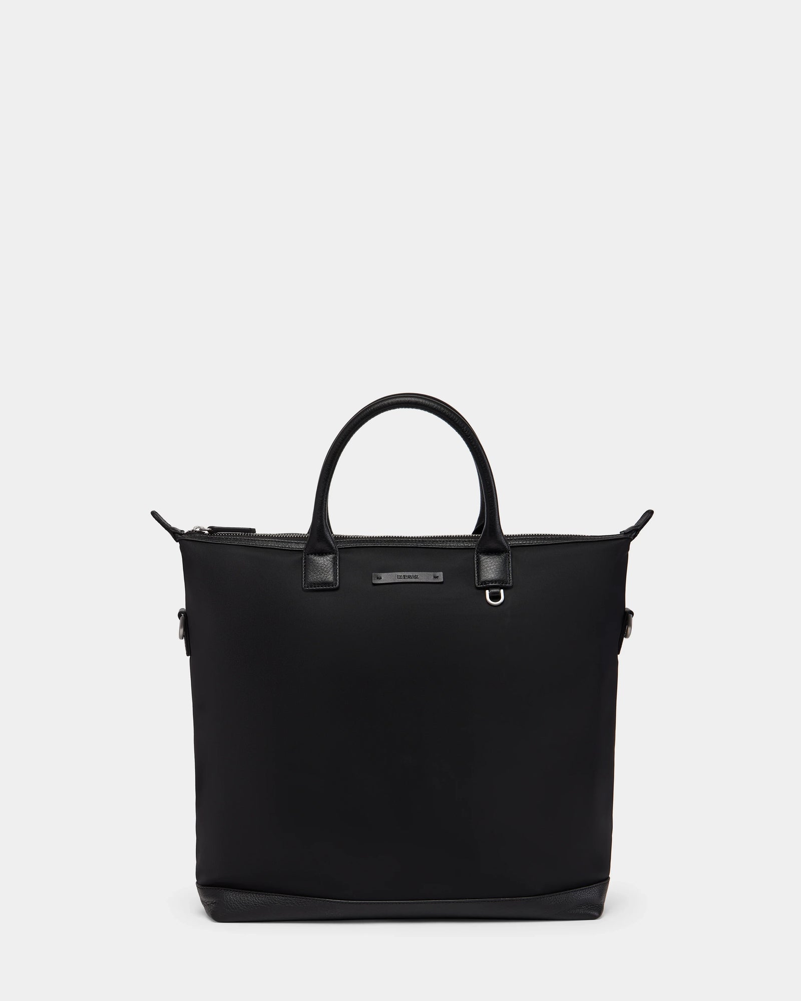 The Rudsak DEXTER - 9324513 Black unisex tote features black canvas, dual handles, a zippered top, and a small metal brand plate on the front, shown against a plain light gray background.