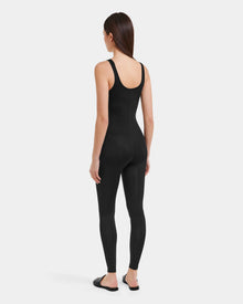 A woman with long brown hair stands facing away, wearing the Rudsak LAWREN - 8523054 Pattern sleeveless, black fitted bodysuit and black flat sandals against a plain light background.