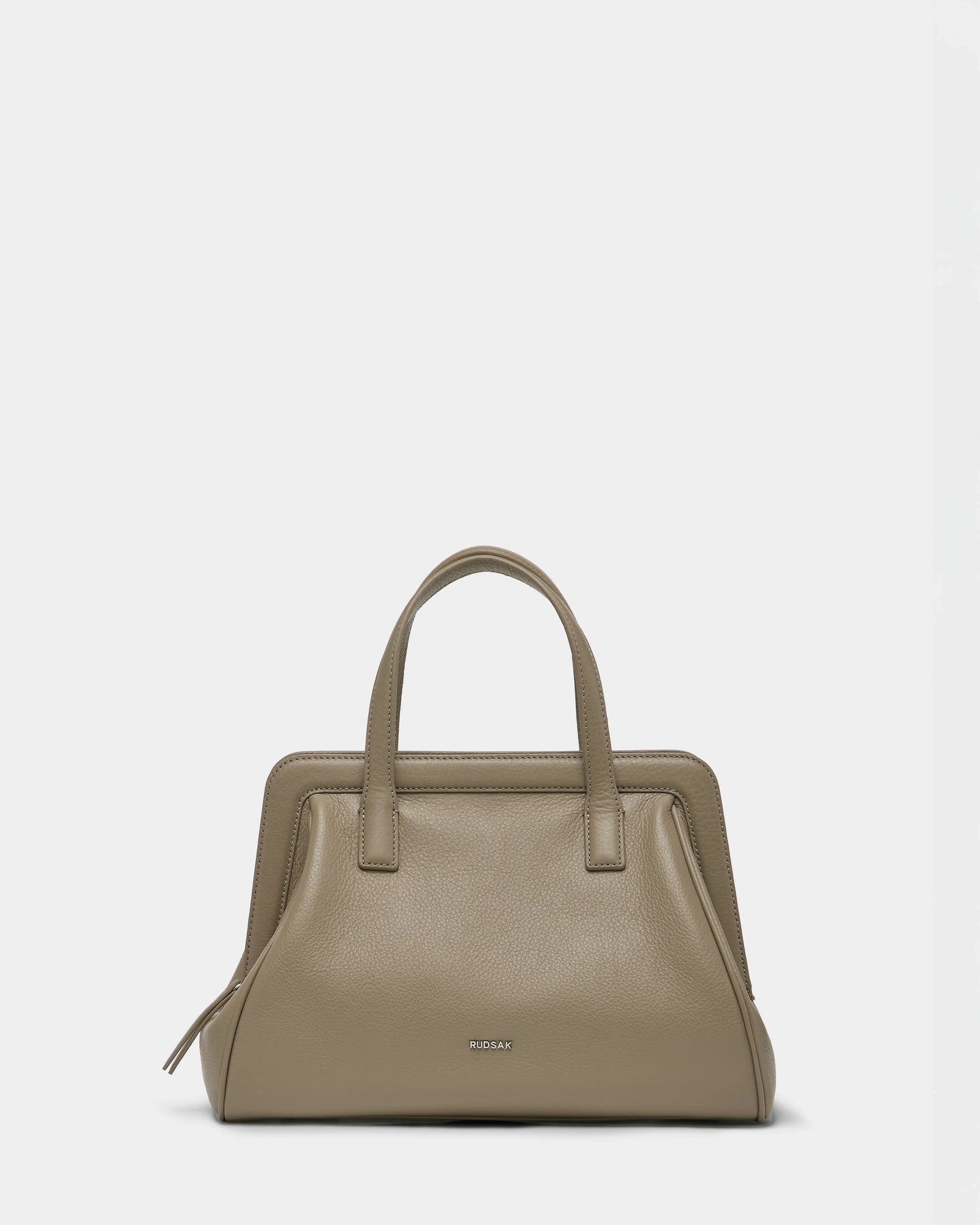 The Rudsak TAULLA - 8325002 Almond is a women’s pebble leather satchel in taupe with two handles, a structured rectangular design, metallic brand lettering on the front, and a spacious interior, shown against a plain white background.