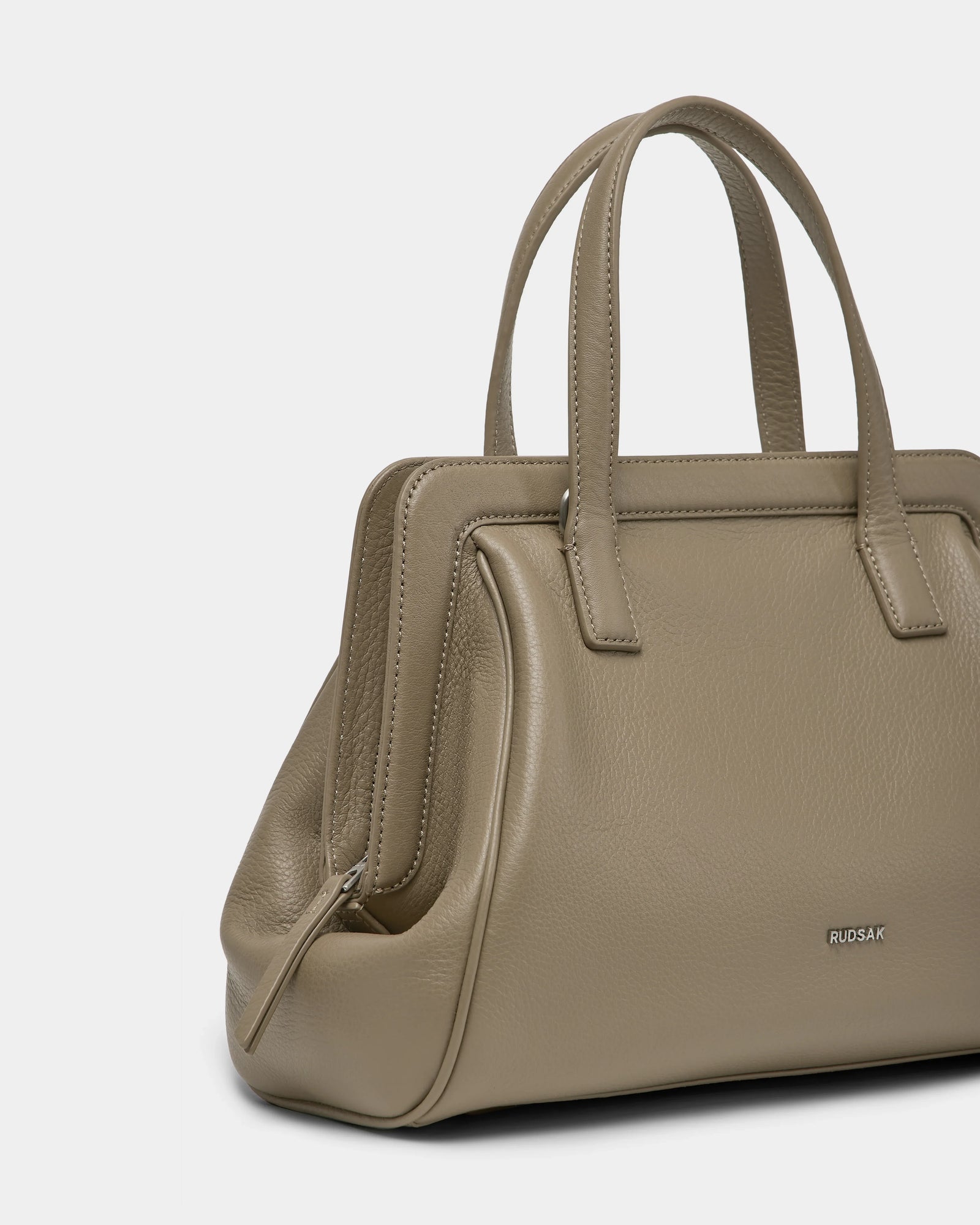 The Rudsak TAULLA - 8325002 Almond is a taupe women’s pebble leather satchel with double handles, a structured rectangular shape, removable shoulder strap, and subtle metallic branding. Stylish and practical, shown on a plain white background.