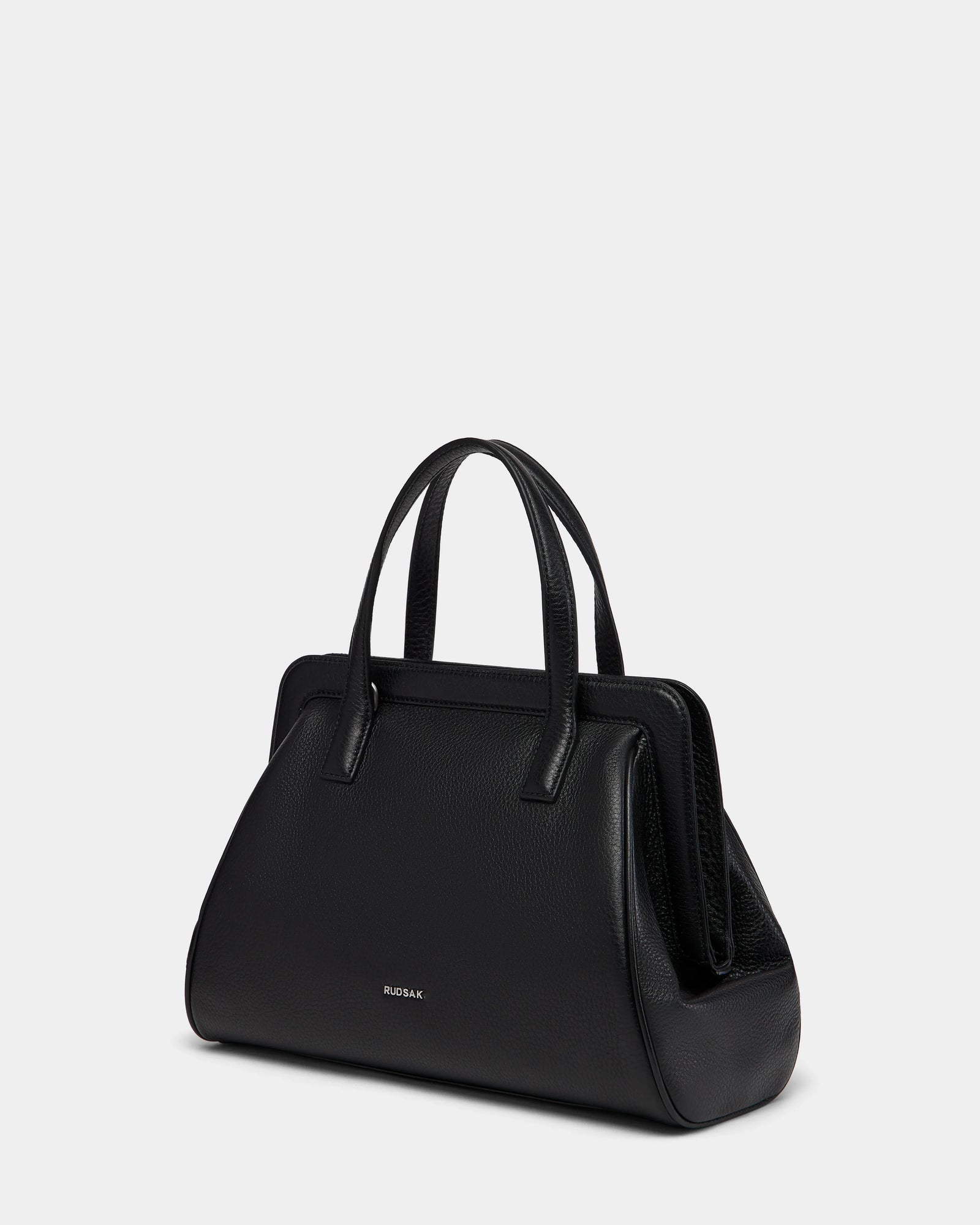 The TAULLA - 8325002 Black by Rudsak is a women’s black pebble leather satchel with two top handles, a structured trapezoidal shape, zippered top closure, and Rudsak embossed in small gold letters on the front against a light gray background.