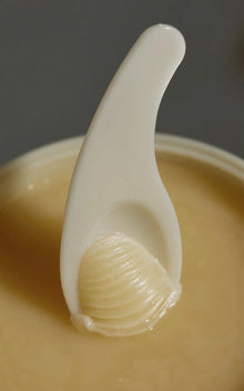 Close-up of The Cleansing Balm on a white spatula showcasing smooth translucent texture with open jar and gray background