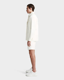 A young man stands in profile against a plain background, wearing the Rudsak KERRY - 9124003 Eggshell M unisex jacket with eco-friendly insulation, paired with textured shorts and sneakers. His hair is slicked back and his arms rest naturally by his sides.