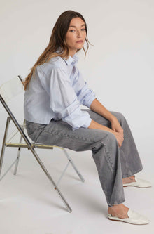 Woman sitting on a chair wearing a light blue shirt and gray pants against a white background