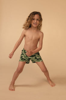Child wearing green and yellow floral swim trunks with a wide stance against a beige background.