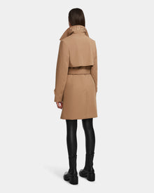 A person faces away in the Rudsak KIMBERLY - 8124801 Camel trench, featuring a tan water-repellent finish, belted waist, vented back, and RUDSAK on the collar. Styled with black leggings and ankle boots against a plain white background.