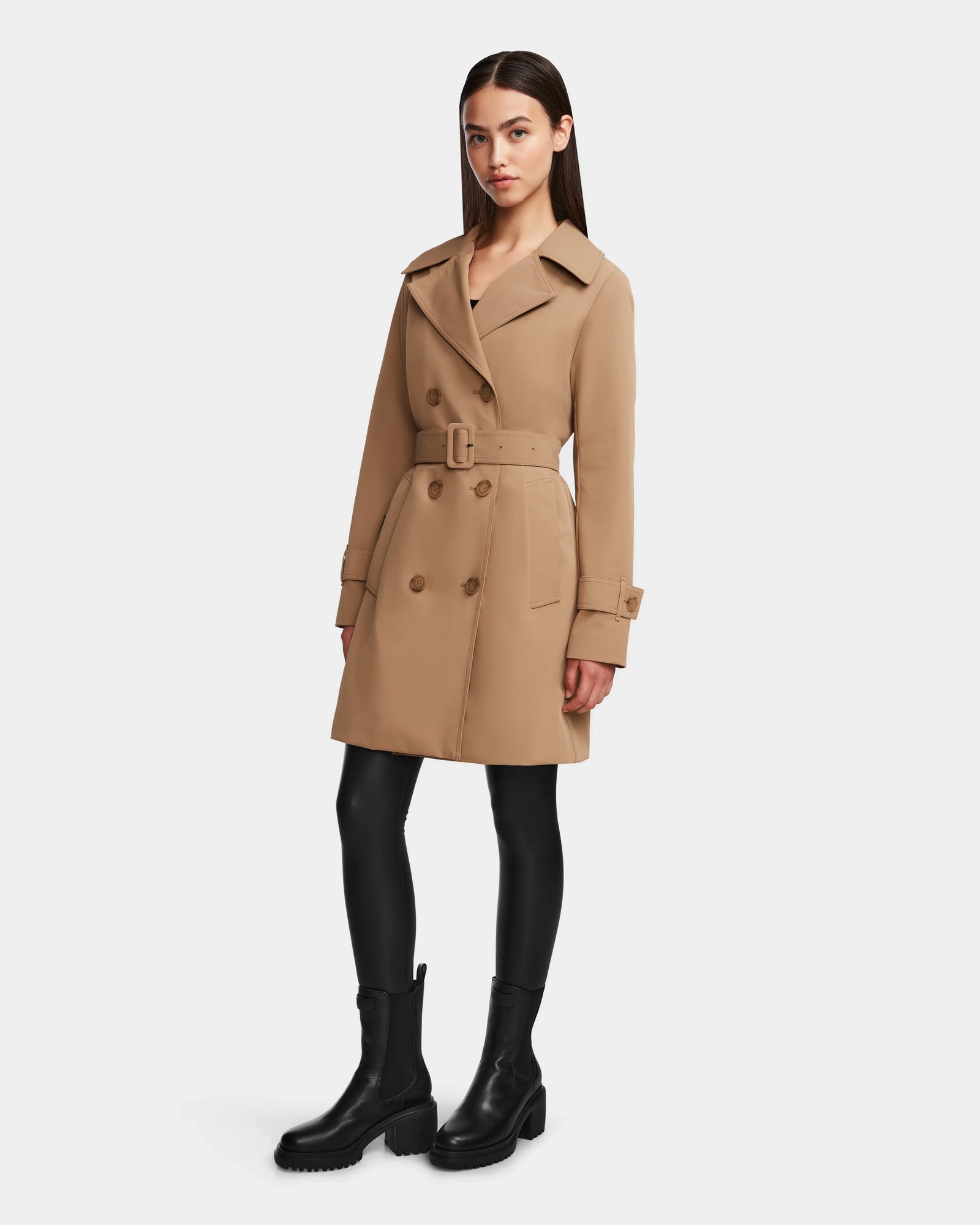 A woman stands against a white background wearing the Rudsak KIMBERLY - 8124801 Camel belted trench coat, paired with black leggings and chunky black boots.