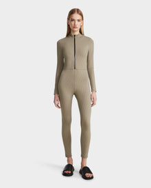 A woman faces forward wearing the Rudsak SHANYA - 8523526 Cement Pattern fitted, long-sleeve zip-up bodysuit in light olive green stretch fabric, paired with black open-toe slide sandals. The background is plain white.