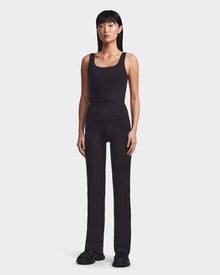 A woman stands facing forward, wearing Rudsaks ADRIANA - 8523550 Black flared-leg leggings with a sleeveless tank top and black sneakers against a plain white background.