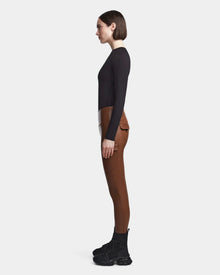 A woman stands in profile against a white background, wearing Rudsak's SIEN L - 8521042 COHIBA: high-waisted brown pants with pockets, styled with a fitted black long-sleeve top and black lace-up boots.