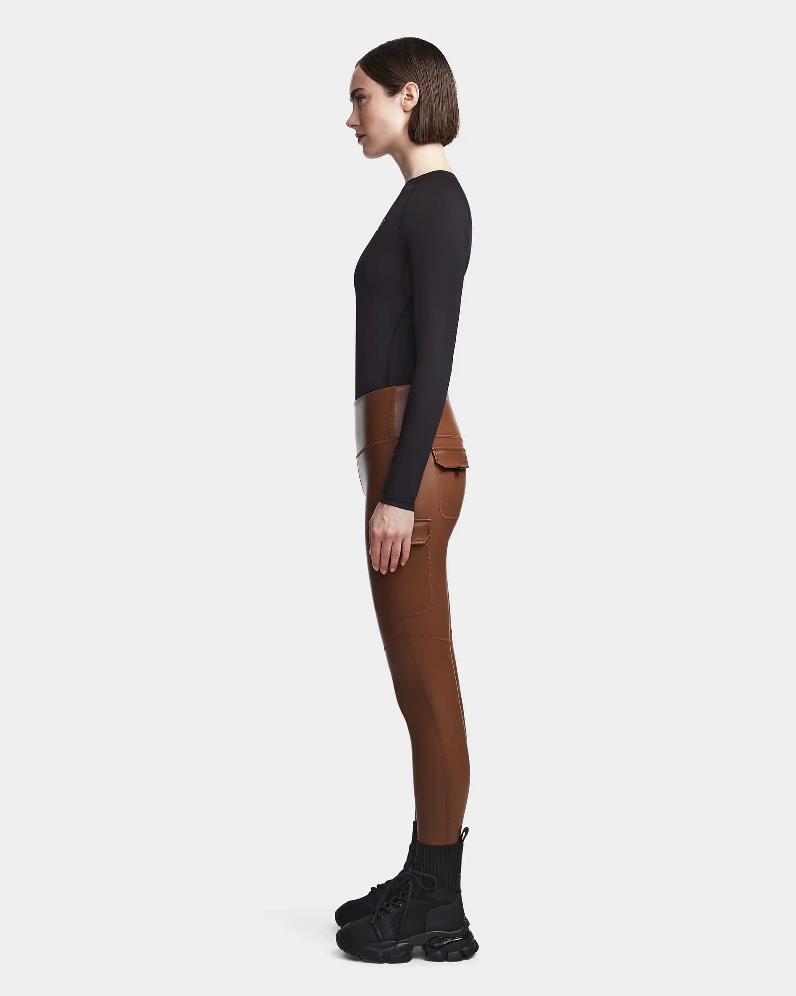 A woman stands in profile against a white background, wearing Rudsak's SIEN L - 8521042 COHIBA: high-waisted brown pants with pockets, styled with a fitted black long-sleeve top and black lace-up boots.