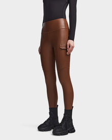 A person wears Rudsak’s SIEN L - 8521042 COHIBA brown vegan leather leggings with a side pocket, styled with a black long-sleeve top, socks, and chunky sneakers against a white background—ideal for highlighting activewear essentials.