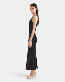 A woman stands in profile wearing the Rudsak WANDA - 8524035 Black maxi dress, crafted from stretch performance fabric and paired with white high-heeled shoes, exuding contemporary elegance against a plain white background.