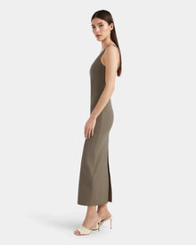 A woman with long straight hair stands sideways, showcasing Rudsaks WANDA - 8524035 Cement maxi dress with a side slit and white heeled sandals, exuding contemporary elegance against a plain white background.