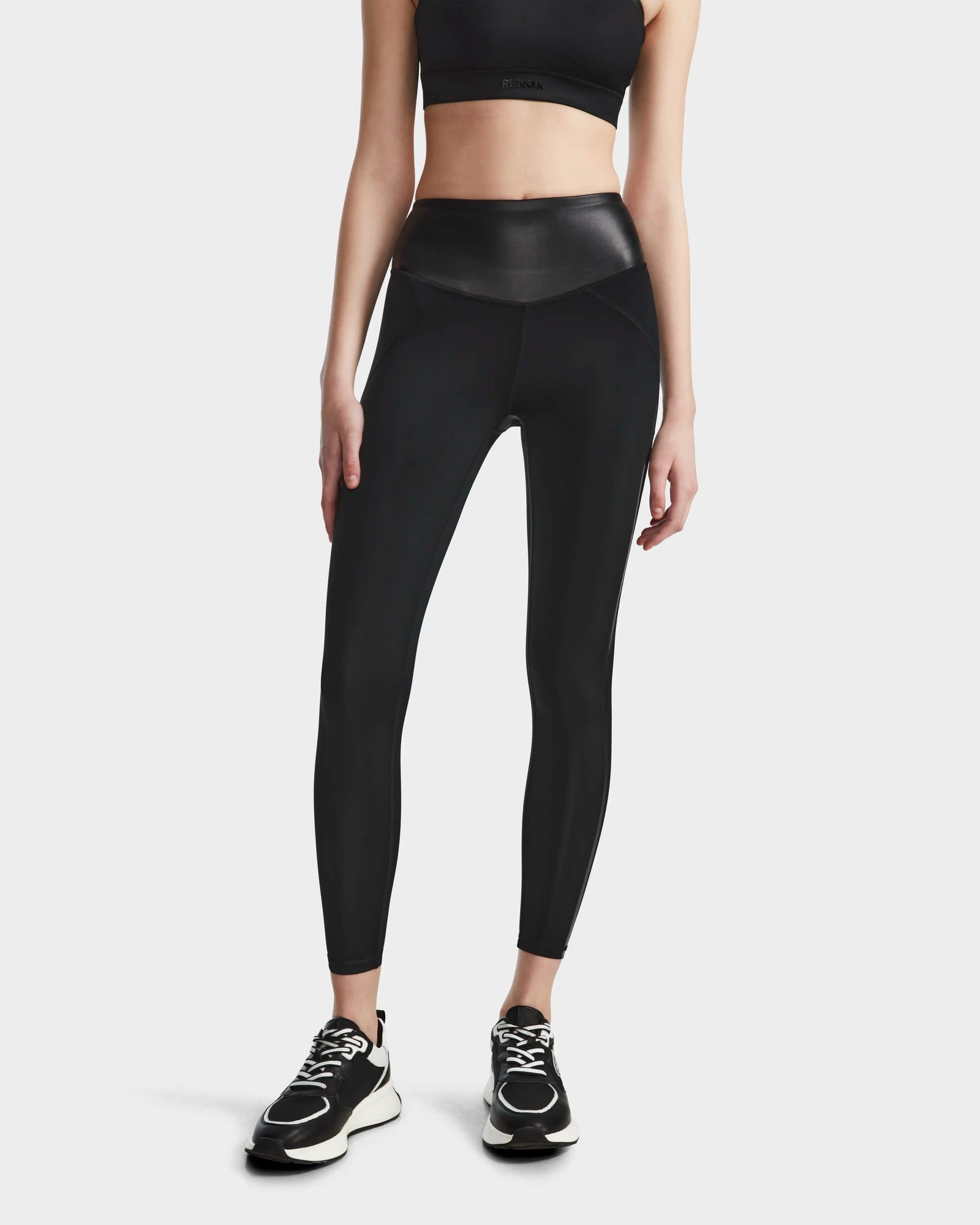 A woman models the Rudsak THEA - 8523003 BLACK high-rise leggings with a vegan leather waistband, paired with a black sports bra and sneakers, against a plain white background. Her arms are by her sides and her head is not visible.