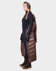 A woman in profile wears Rudsak’s ELSIE - L121525 CAOCAO women’s maxi down puffer vest with RDS-certified down, a black bodysuit, black gloves, brown earmuffs, and brown lace-up boots against a plain light background.