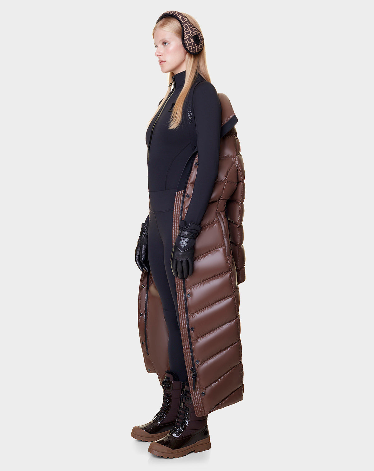 A woman in profile wears Rudsak’s ELSIE - L121525 CAOCAO women’s maxi down puffer vest with RDS-certified down, a black bodysuit, black gloves, brown earmuffs, and brown lace-up boots against a plain light background.