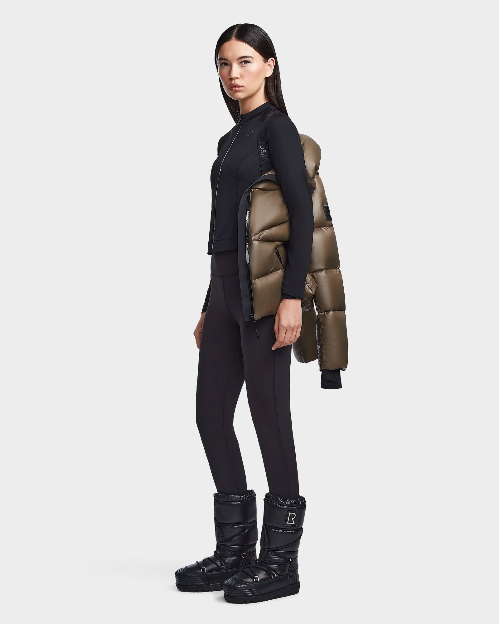 A woman stands sideways against a white background, draping the Rudsak CHRISTELLE S - L123524 Light Olive women’s chevron quilted down puffer jacket over one shoulder while wearing black boots, leggings, and a fitted zip-up top.