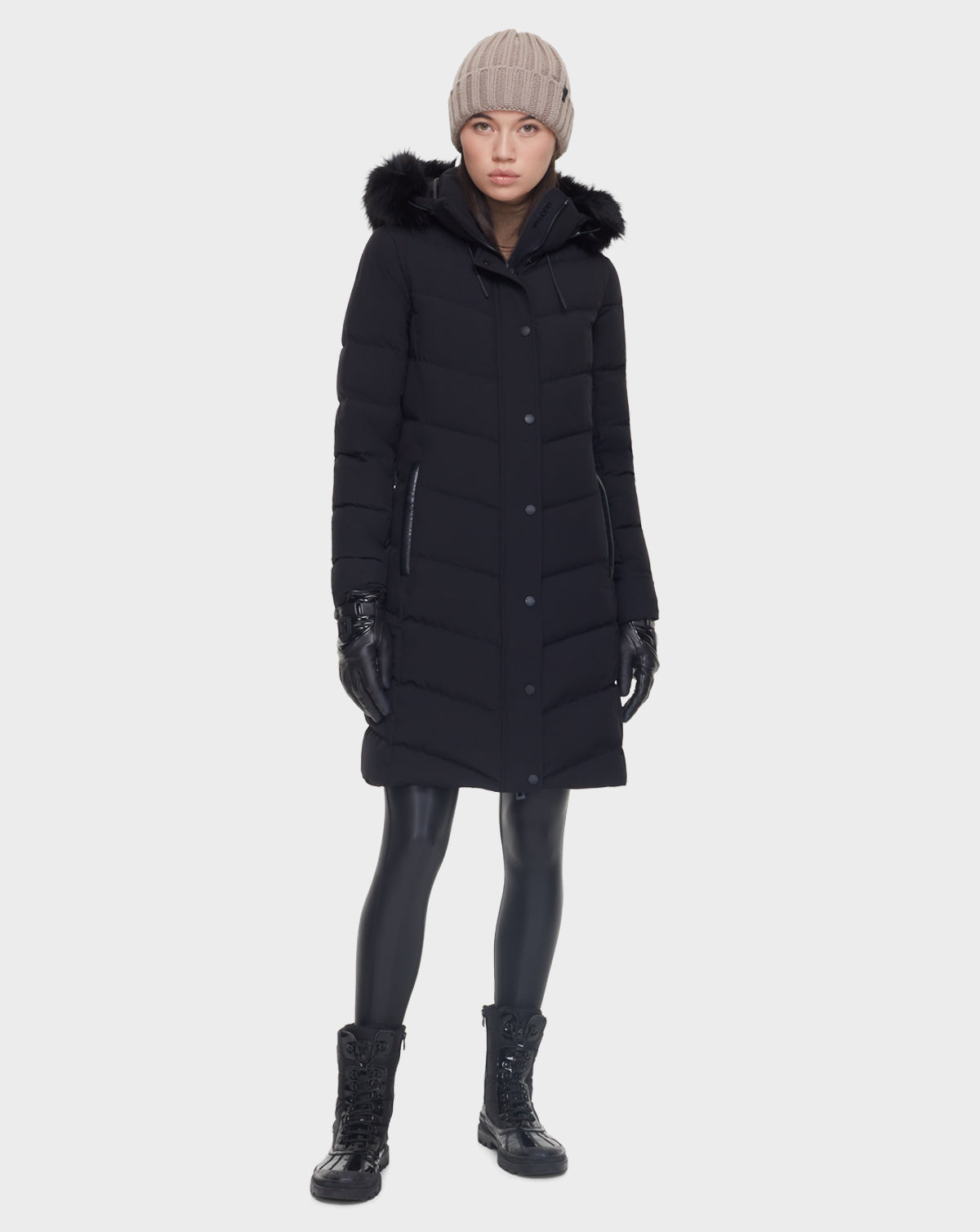 A person wearing the Rudsak ELISA - L121512 BLACK quilted down puffer with faux fur hood, black gloves, shiny leggings, lace-up boots, and a beige knit beanie stands against a plain white background.