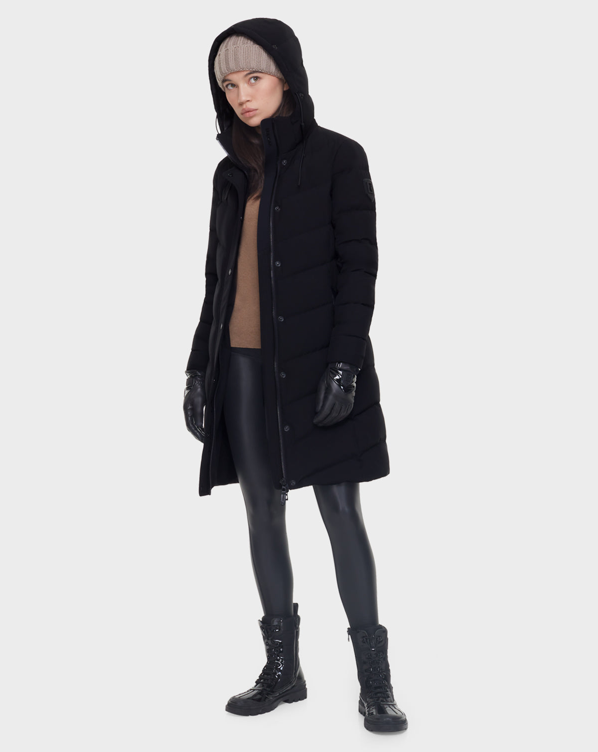 A woman wears the Rudsak ELISA - L121512 BLACK quilted down puffer coat with a tan sweater, black leggings, lace-up black boots, black gloves, and a beige knit beanie against a white background.