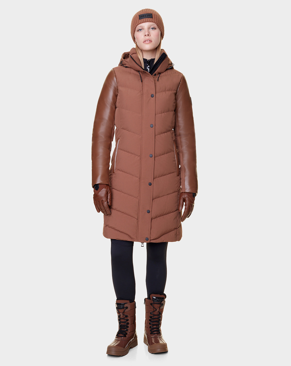 A person wears the Rudsak ELSA - L121519 TAUPE women’s quilted down puffer coat with leather sleeves, plus brown gloves, boots, a knit beanie, and black leggings—showcasing sustainable design—against a plain white background.