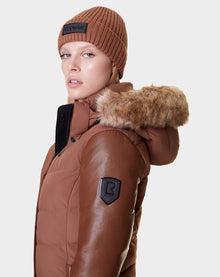 A woman in the Rudsak ELSA - L121519 TAUPE quilted down puffer with faux fur hood and brown beanie faces the camera against a light gray background. The jacket features sustainable design, a black sleeve patch, and black fastenings.