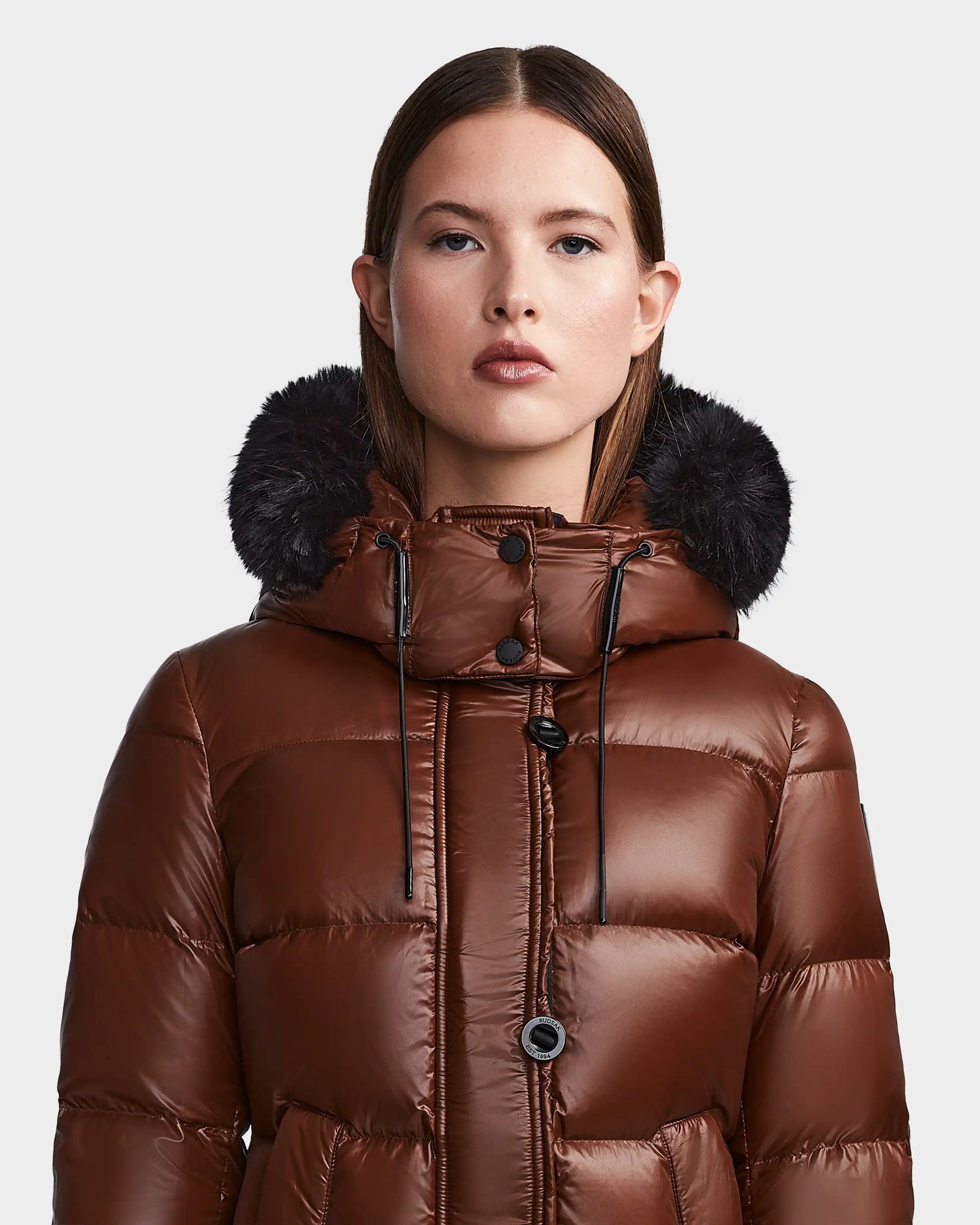 A woman with straight brown hair wears the shiny brown Rudsak MEYA S - L123521 Cohiba down puffer jacket with a removable black faux fur-trimmed hood, standing against a plain light gray background.