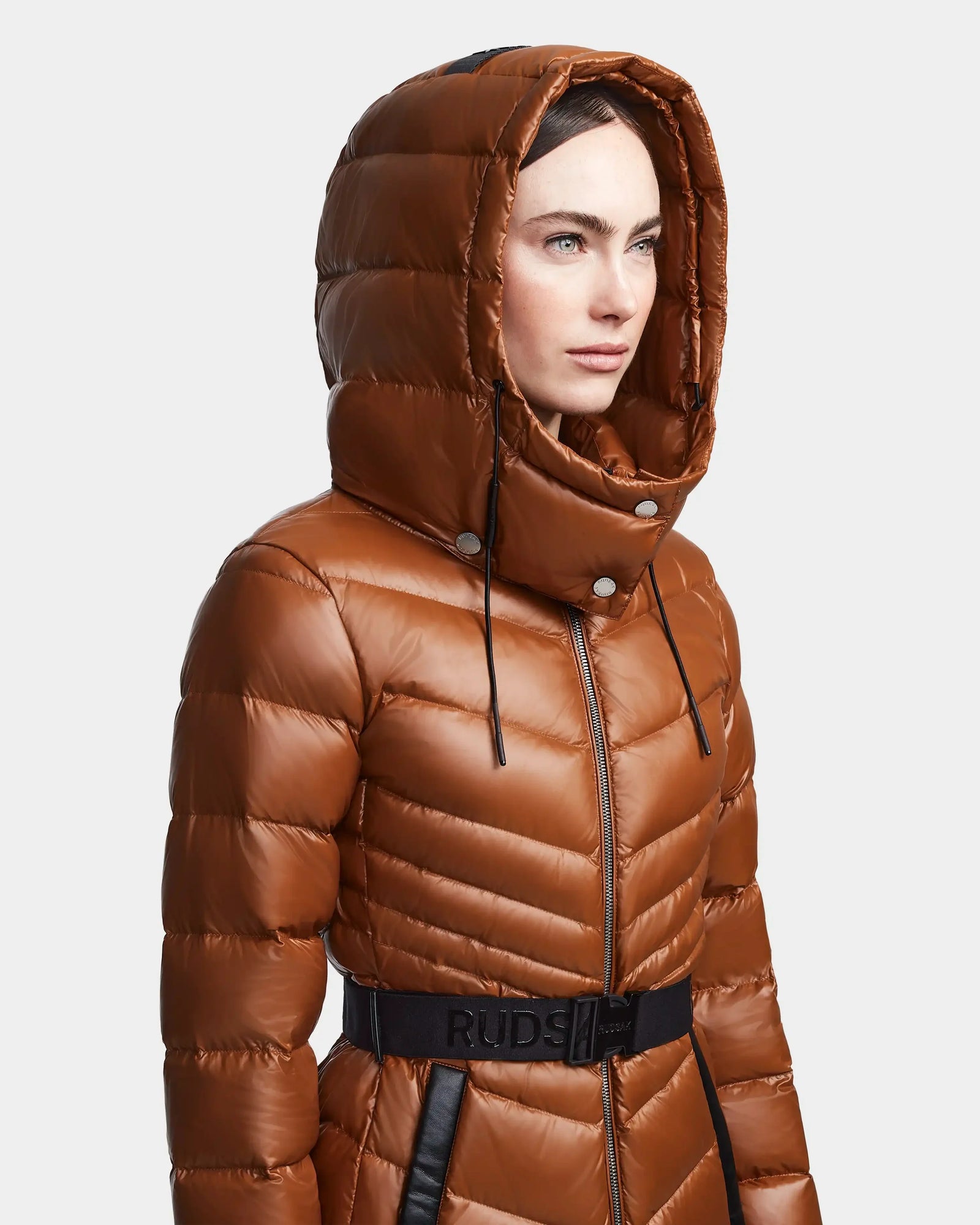 A woman stands against a plain light background, looking to the side, wearing the Rudsak MALAYA - L123510 Dark Camel women’s down puffer jacket featuring a large hood, black belt, and subtle herringbone quilting.