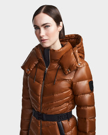 A woman wearing the Rudsak MALAYA - L123510 Dark Camel down puffer jacket with herringbone quilting and a black belt stands against a plain light gray background.