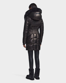 A person stands facing away, wearing the Rudsak SHAUNA X - 8121568 BLACK—a shiny black puffer with a large fur-trimmed hood—paired with black leggings and boots against a white background; a chic women's winter coat for cold days.