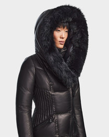 A woman in the Rudsak SHAUNA X - 8121568 BLACK puffer with a large, fur-lined hood stands slightly turned against a plain light background.
