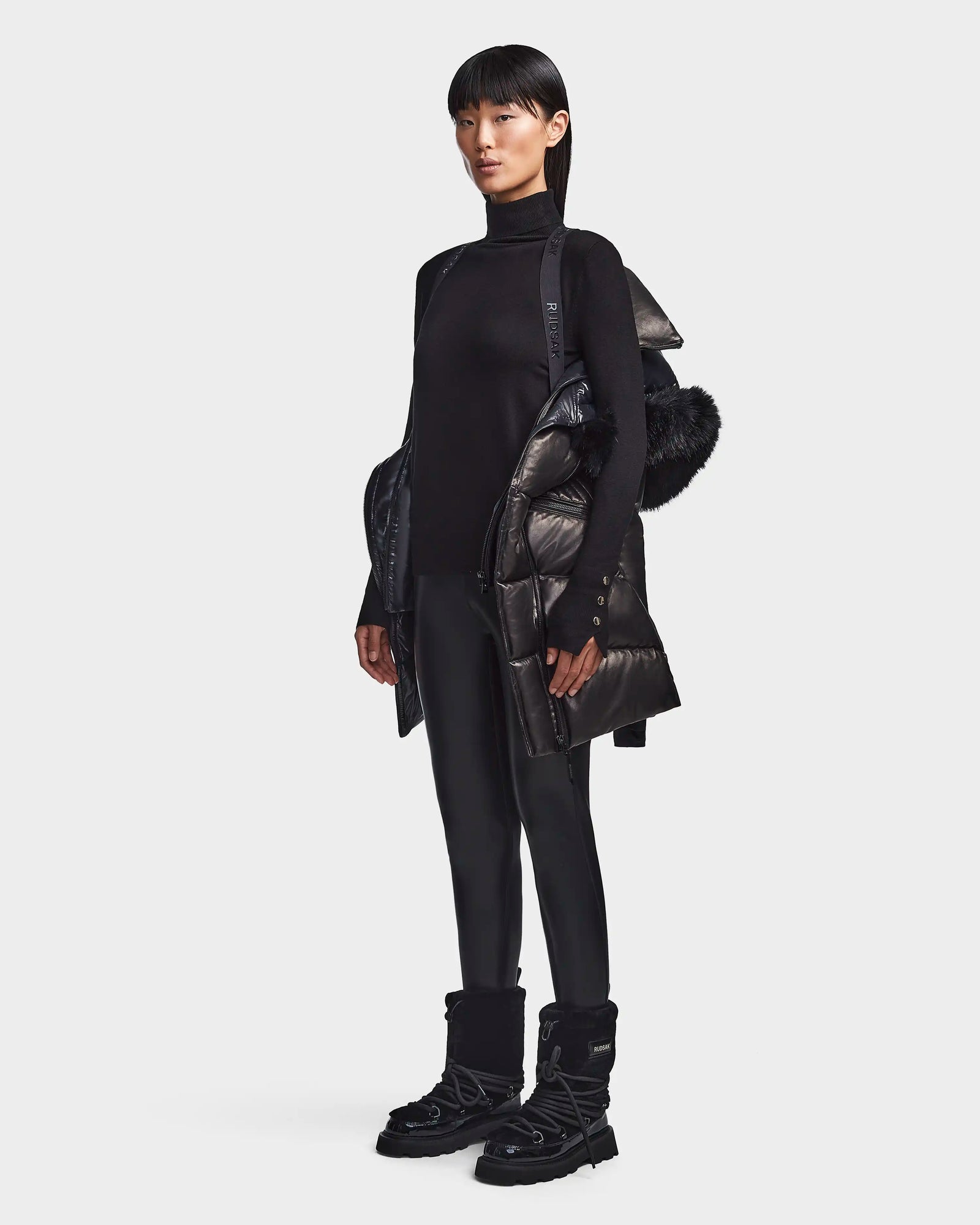 A person wearing the Rudsak SHAUNA X - 8121568 BLACK Cloud leather puffer with fur trim, black turtleneck, pants, and winter boots stands facing forward against a plain light background.