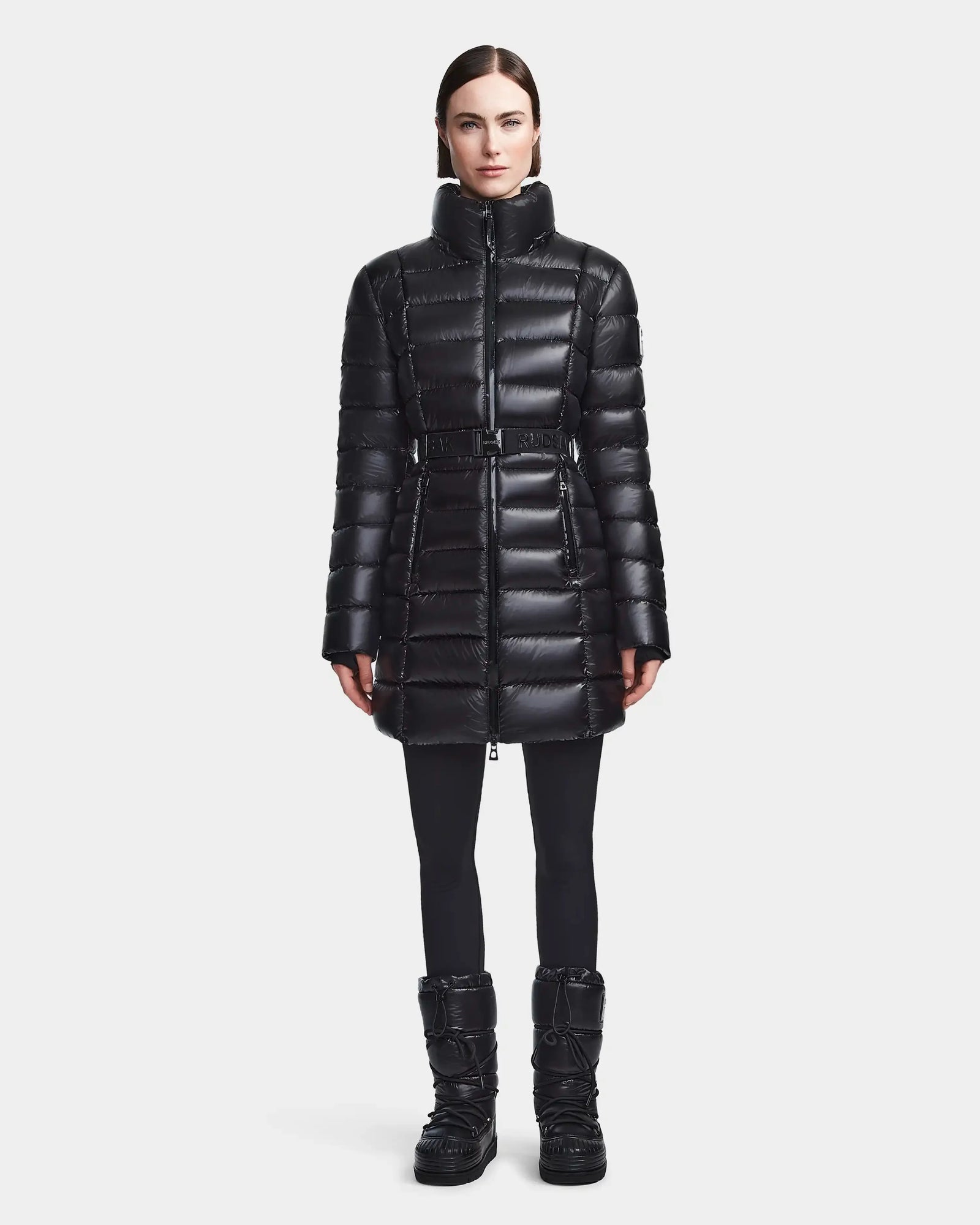 A woman faces forward wearing the Rudsak FRIDA S - L123513 Black mid-length down jacket with a removable hood, paired with black leggings and winter boots, against a plain white background.