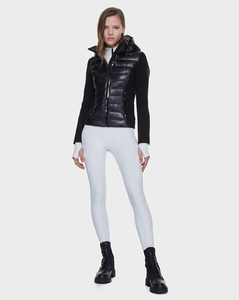 A woman faces forward, wearing the Rudsak MABS - 8121012 BLACK puffer vest filled with RDS-certified down over a white top and leggings. She pairs it with black lace-up boots and stands against a plain white background.