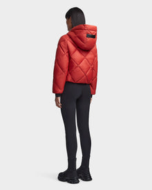 A person stands facing away, wearing the Rudsak BALA - 8123614 Orange reversible hooded jacket with black leggings and chunky black boots against a plain white background.
