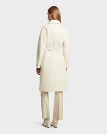 A woman with straight light brown hair models the Rudsak KOURT W - 8124702 Iceland: a long, cream belted wool coat over matching trousers and heeled boots, standing back to camera against a plain light gray background.