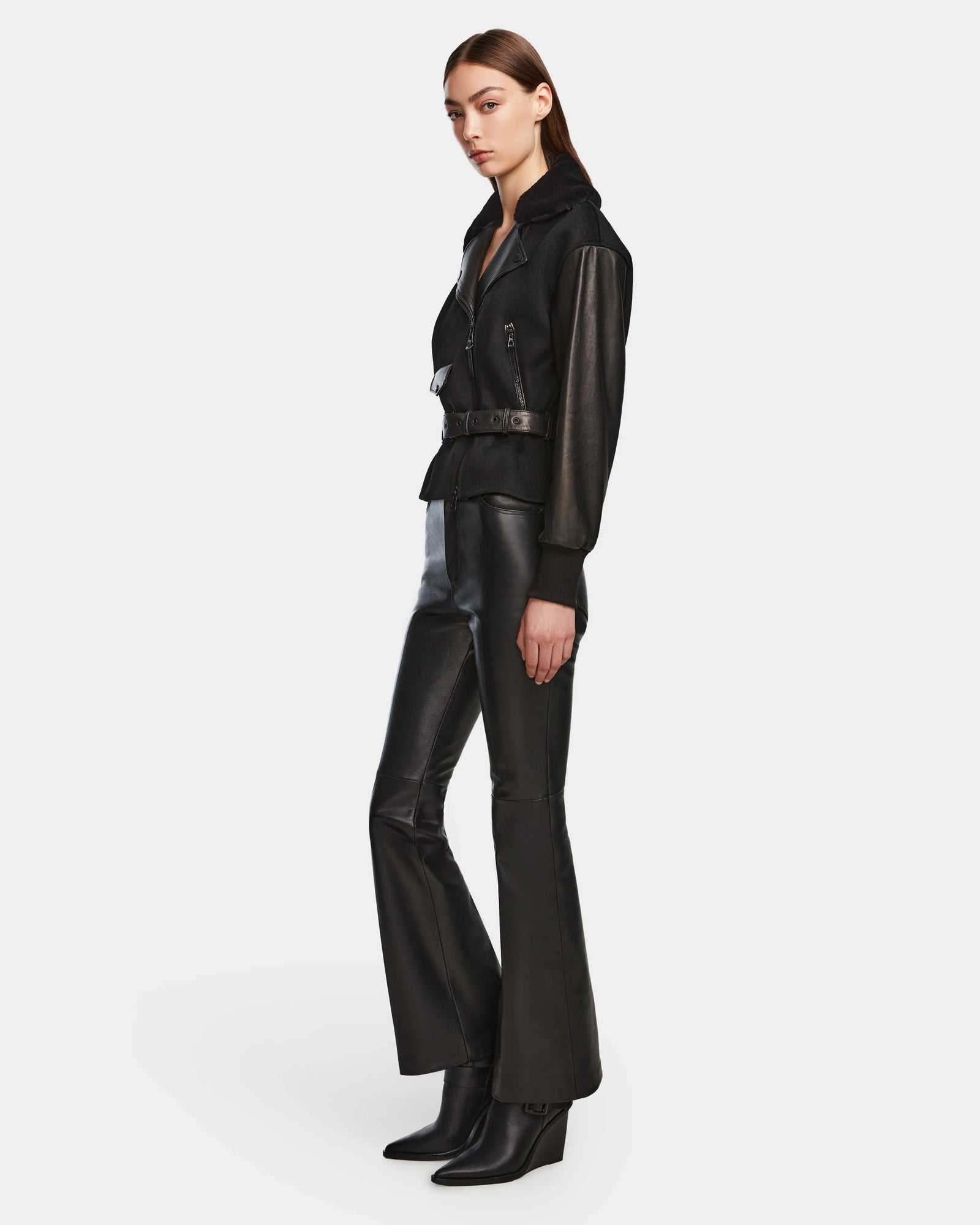 A woman stands in a three-quarter pose wearing the Rudsak HALA - 8124521 Black belted jacket with black leather flared pants and heeled boots, perfect as a chic fall look, against a plain white background.